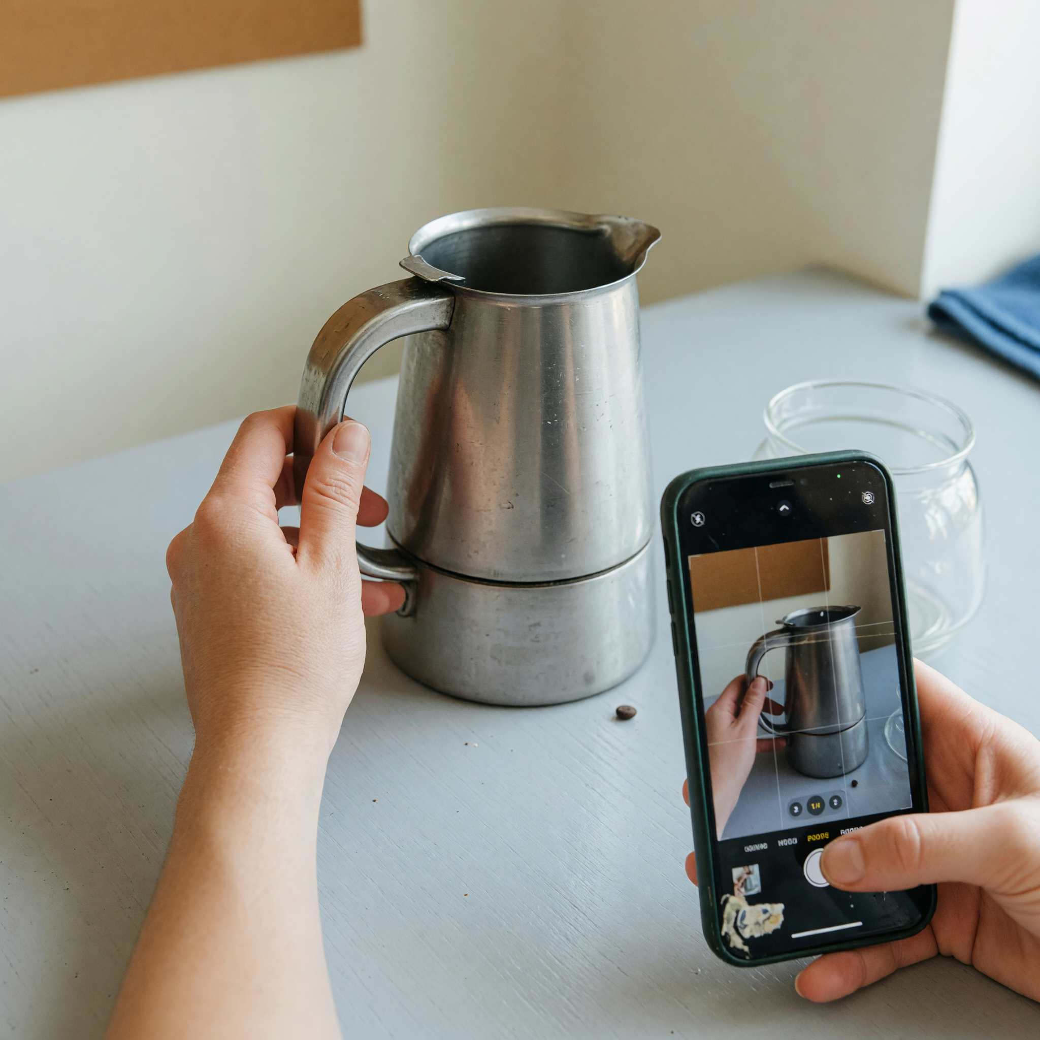 Hands positioning a clean used coffee maker on a neutral table while a phone frames the first listing photo