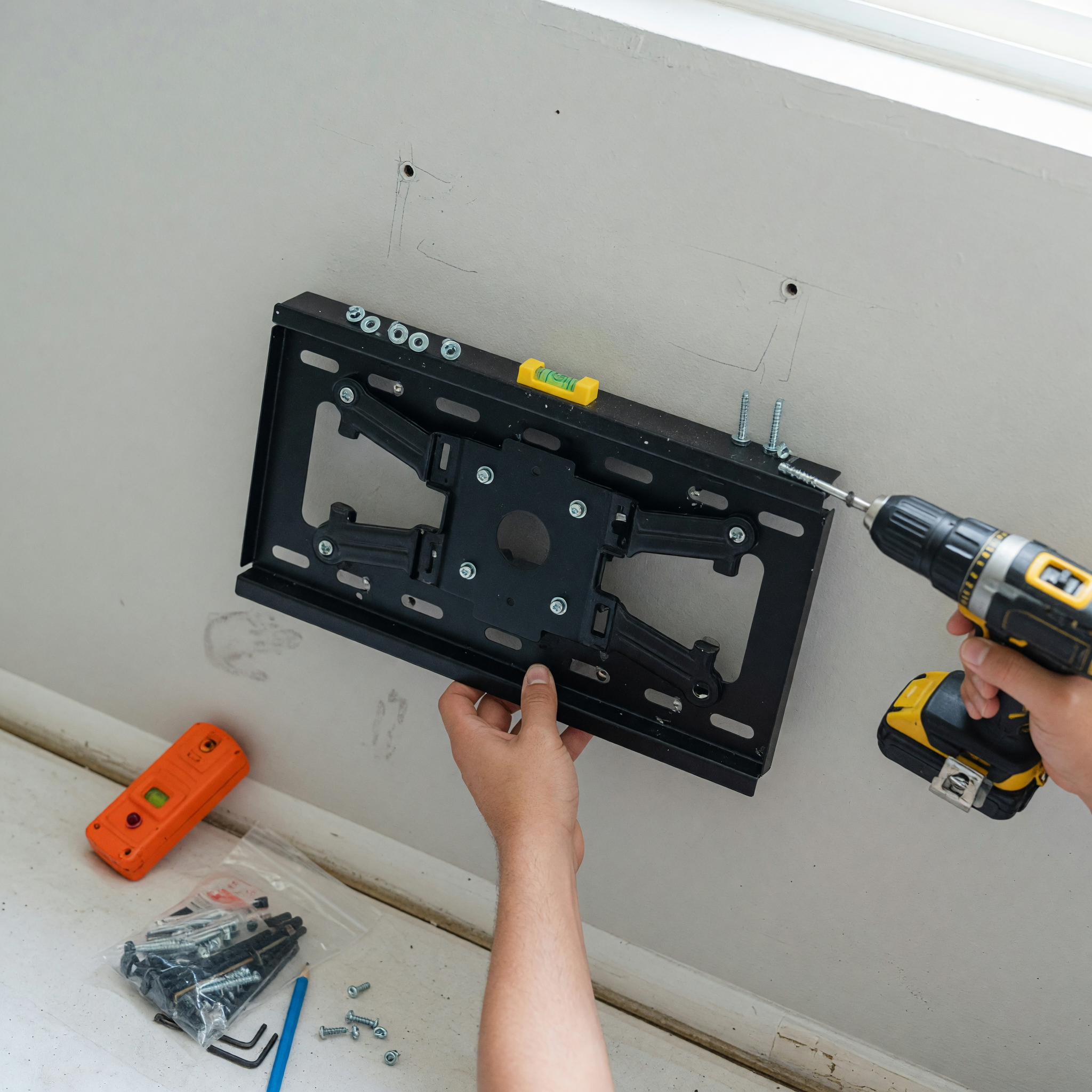 Close view of hands using a cordless drill to secure a black TV wall-mount bracket, with a small bubble level on the bracket showing it is straight