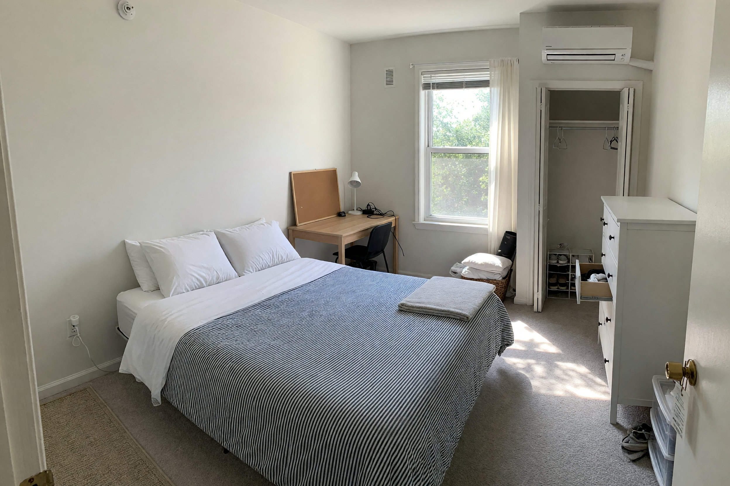 A bright, tidy furnished spare bedroom in a shared US apartment with a made bed, clean linens, a desk, and a sunlit window, ready to rent out