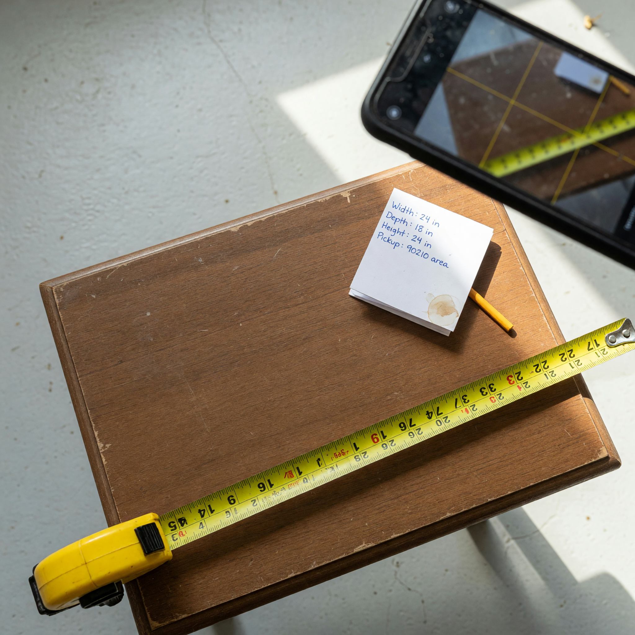 A used side table being measured with a tape measure across its width while a smartphone photographs it in daylight, next to a handwritten card listing dimensions and a pickup area